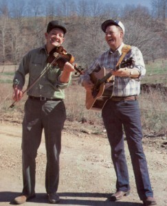 Two men standing on a gravel road, holding a fiddle and a guitar, laughing