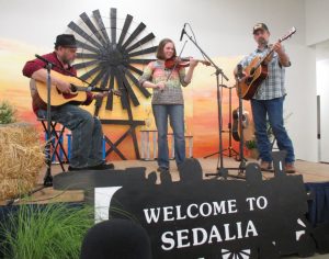 A woman plays fiddle on a stage while two men play guitar