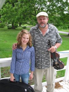 A man and a young woman holding fiddles on the deck of a house