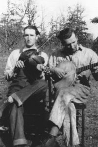 A historic photo of two men, one playing fiddle, one playing banjo, in a field