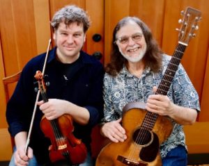 Kristian Bugge holds a fiddle while WB Reid holds a guitar while seated on a bench